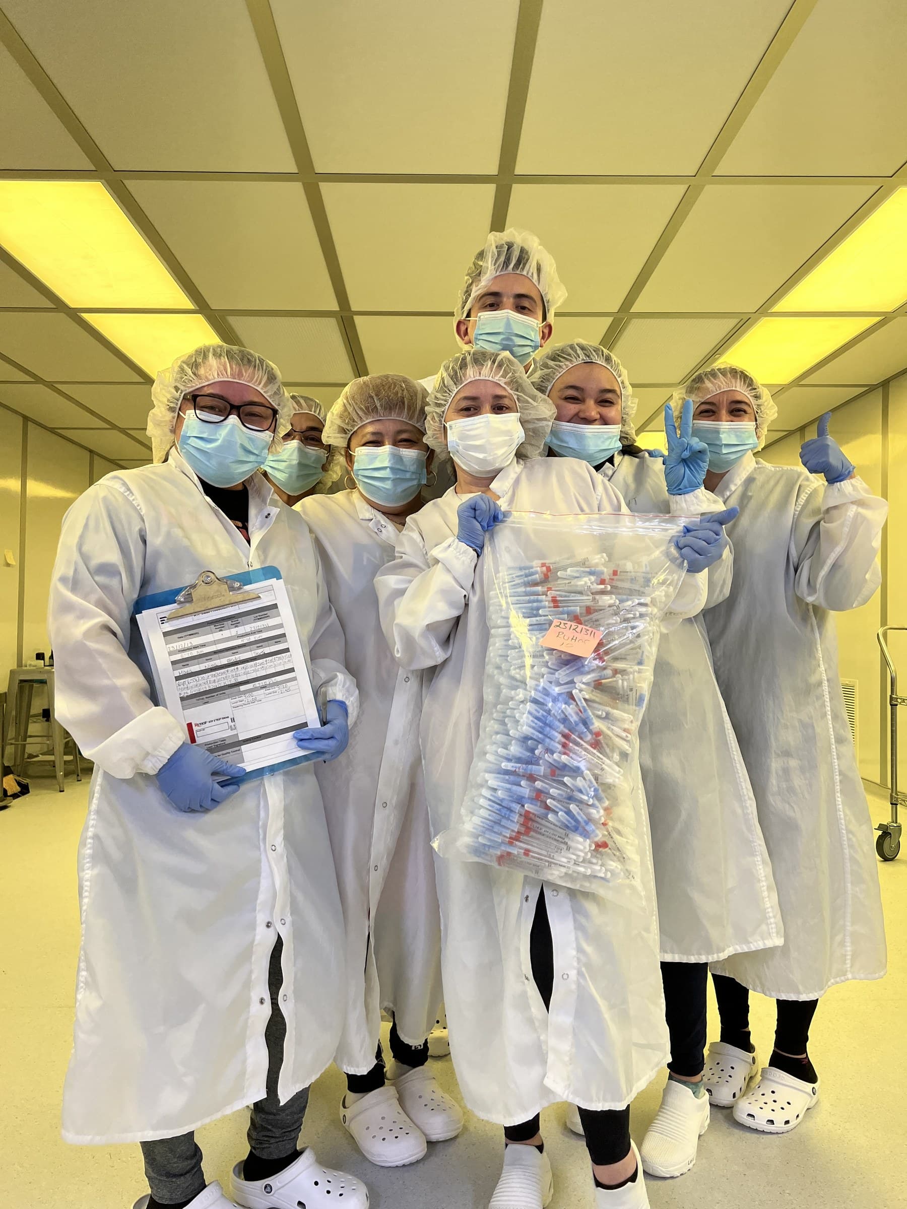 Colleagues in lab coats, masks, and gloves holding a bag of assembled swabs.