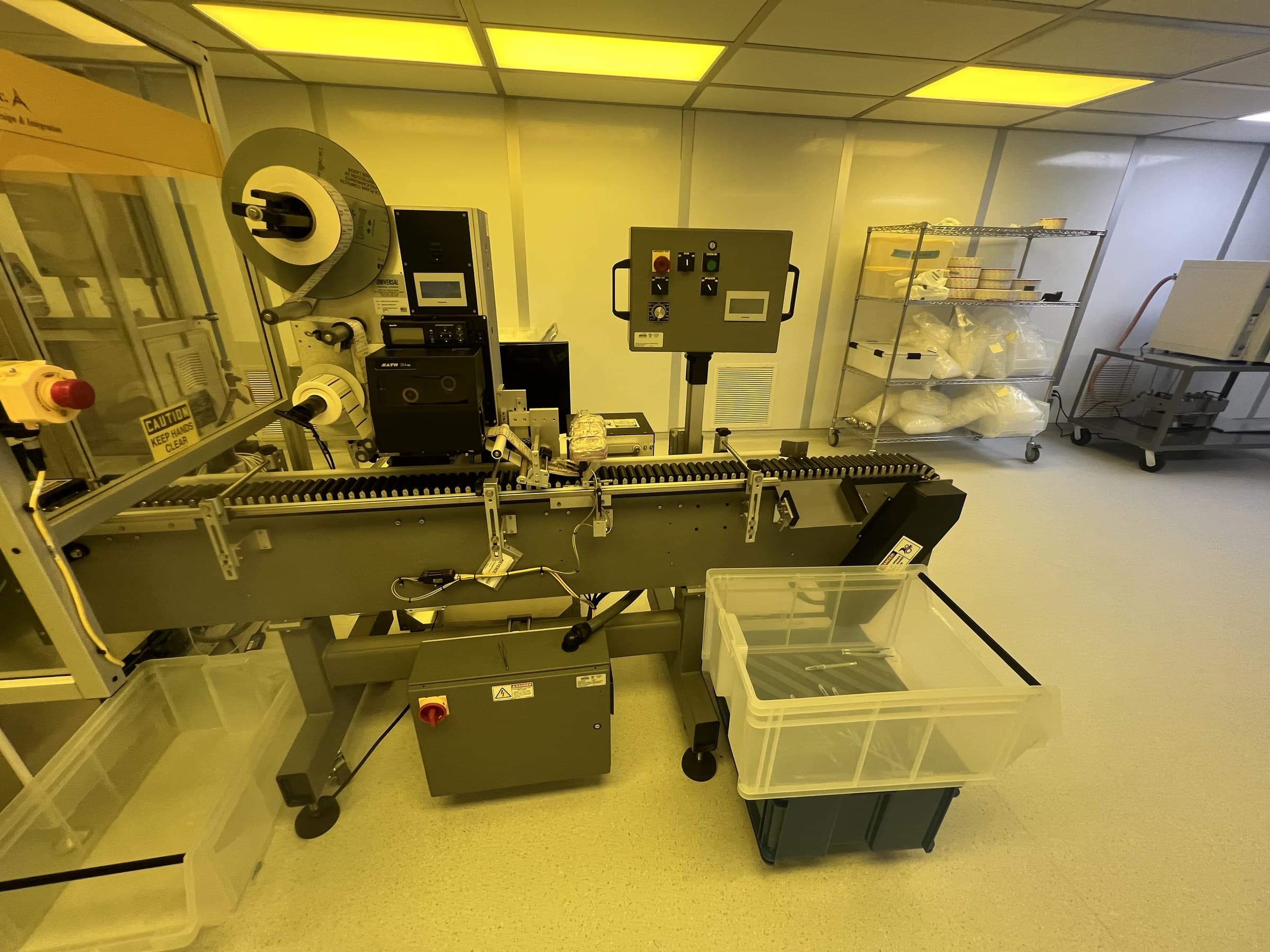 Labeling equipment and a control panel beside a conveyor and collection bin.